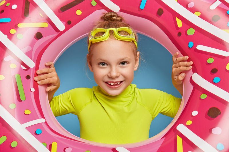 Usměvavá dívka s plaveckými brýlemi se dívá skrz nafukovací kruh ve tvaru růžového donutu, symbol kreativního marketingu a výrazného positioningu produktu.