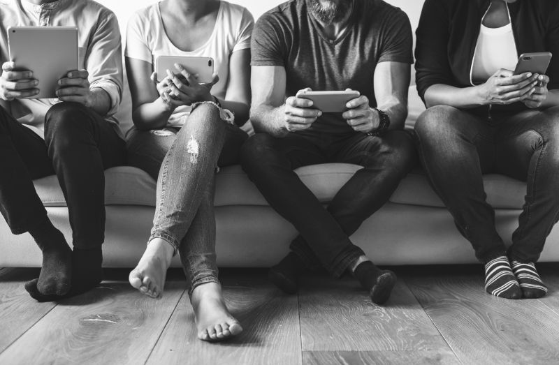 Skupina lidí sedí vedle sebe na gauči a používá chytré telefony a tablet, symbol online komunikace a digitálního marketingu.