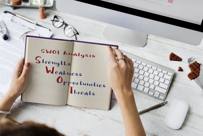 Zápisník s nápisem SWOT analýza a vypsanými body Strengths, Weaknesses, Opportunities, Threats na pracovním stole s klávesnicí a počítačem.